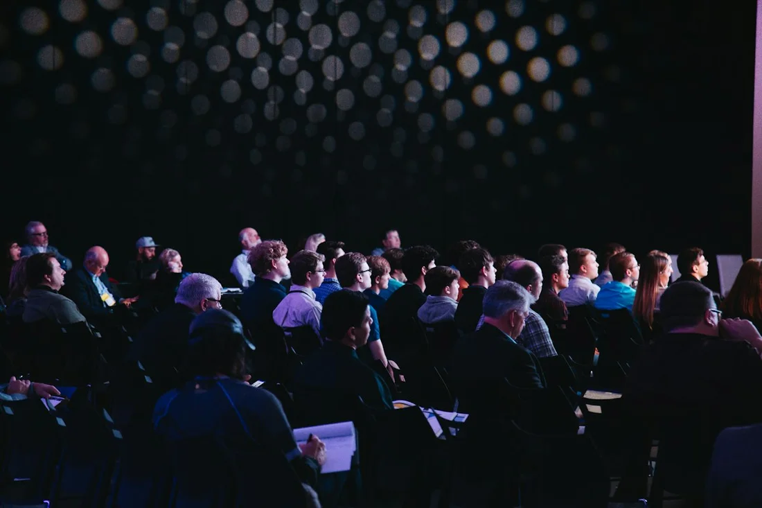 An audience sits in a darkened auditorium facing a lit stage.