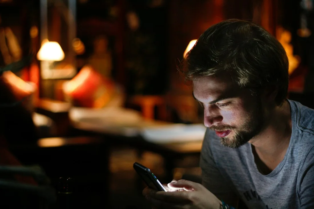 A young person sits alone in a dark room, face lit by a smartphone screen.